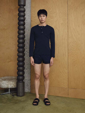 Man wearing a navy long-sleeve shirt and shorts standing against a wooden wall.