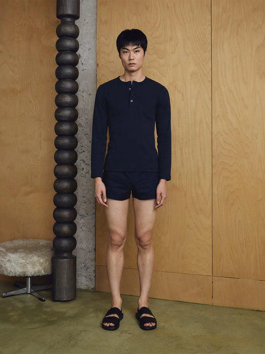 Man wearing a navy long-sleeve shirt and shorts standing against a wooden wall.