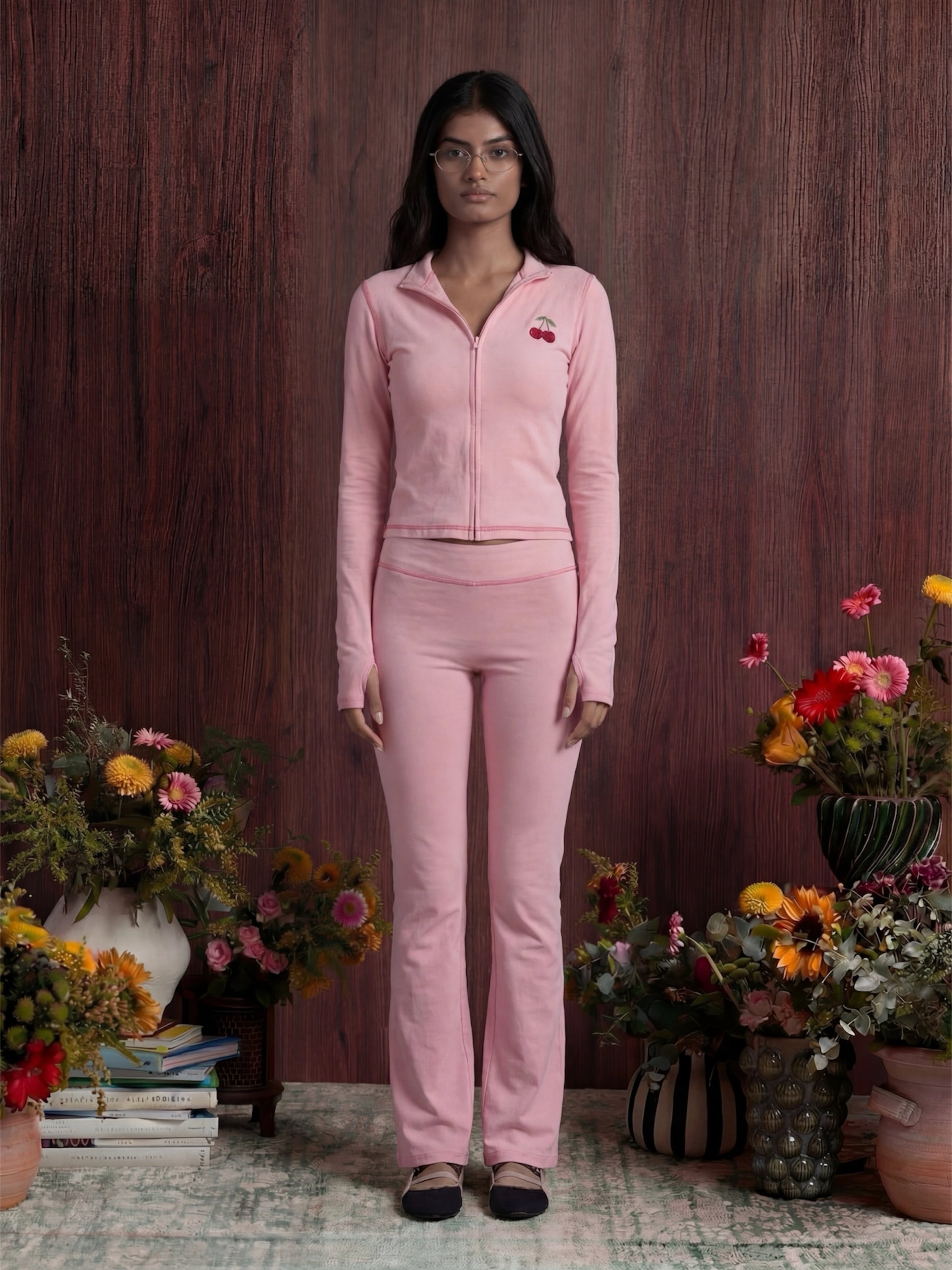 Woman in a pink outfit standing among floral arrangements against a wooden wall.