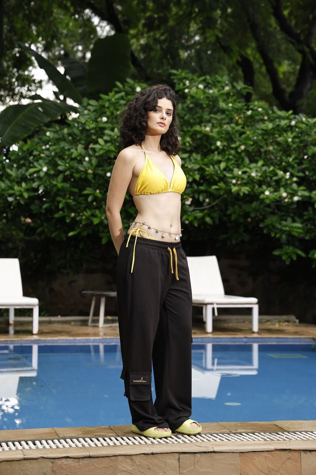 Woman in yellow top and black pants standing by a pool with greenery in the background