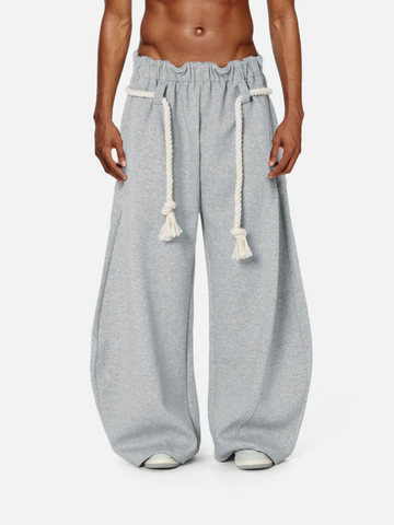 A male model wearing gray baggy barrel sweatpants with a wide-leg fit and elasticized waistband, featuring a rope drawstring.