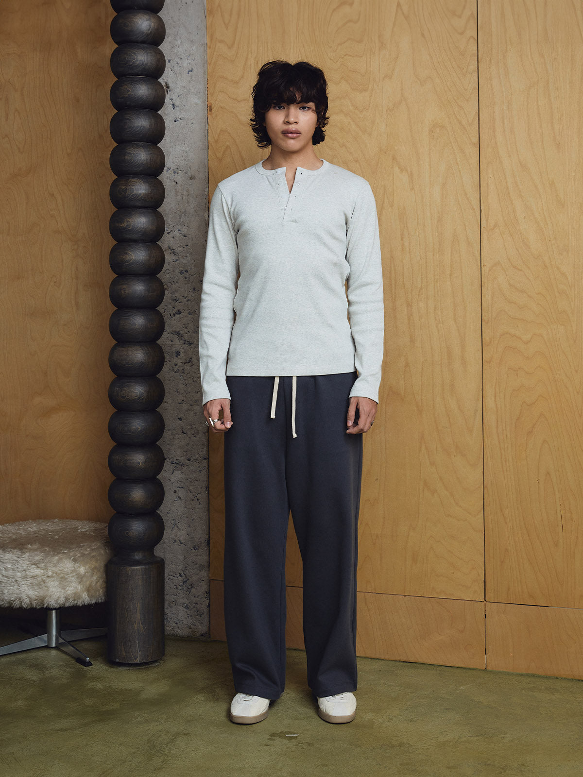 Person wearing a light grey sweater and dark pants standing against a wooden wall.