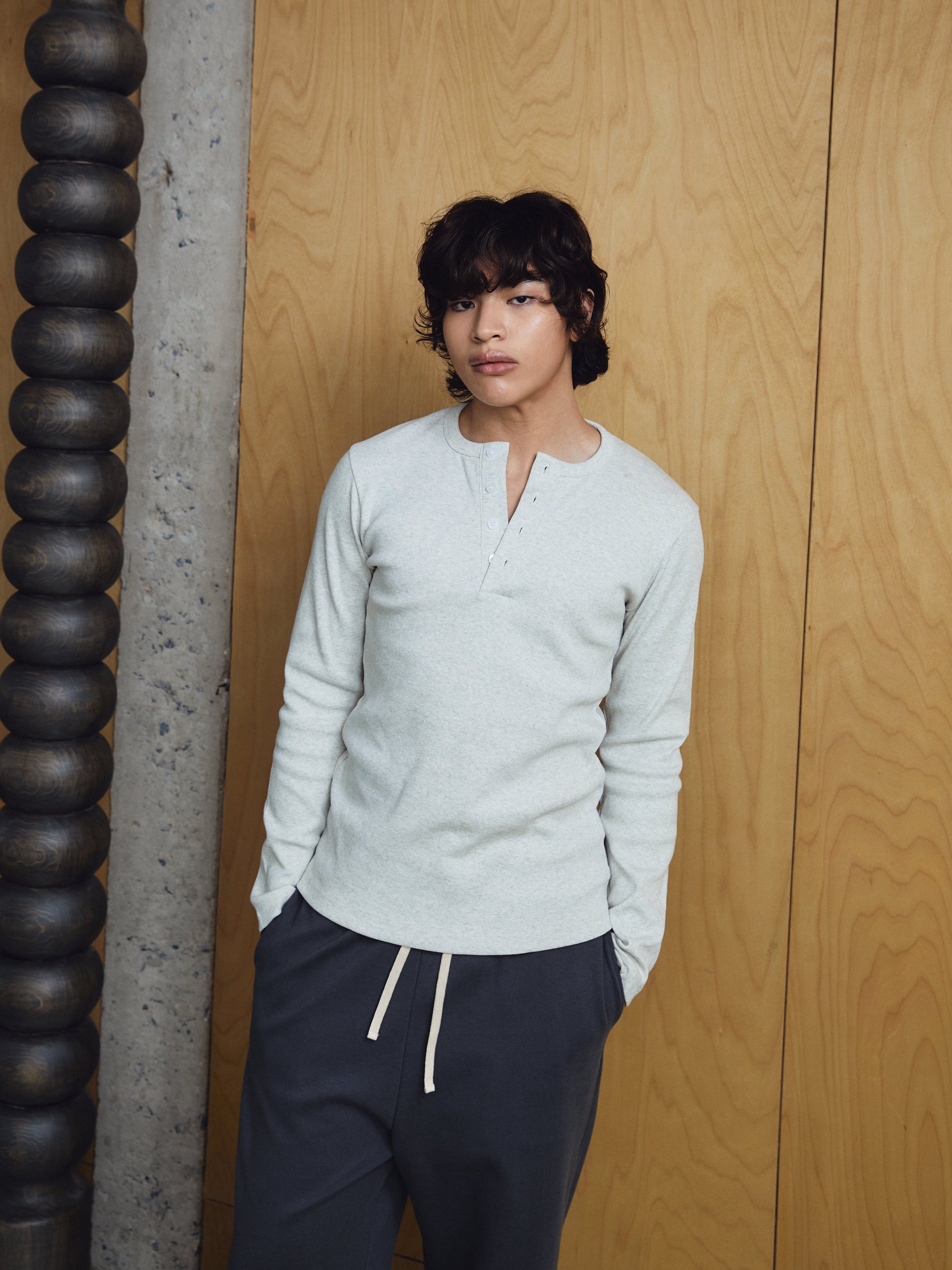 Person wearing a light grey sweater and dark pants against a wooden wall.
