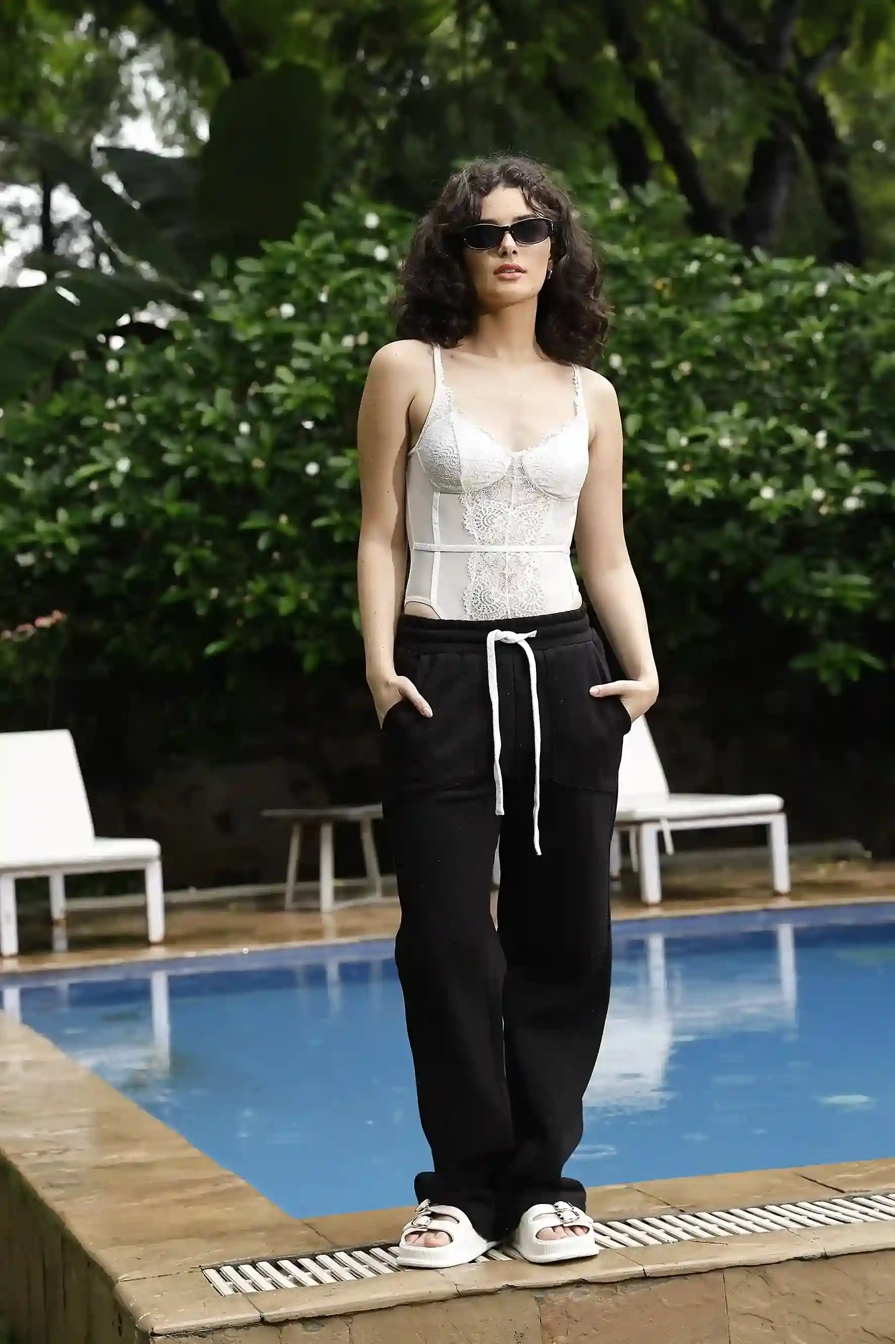 Woman standing by a pool wearing a white top and black pants with sunglasses.