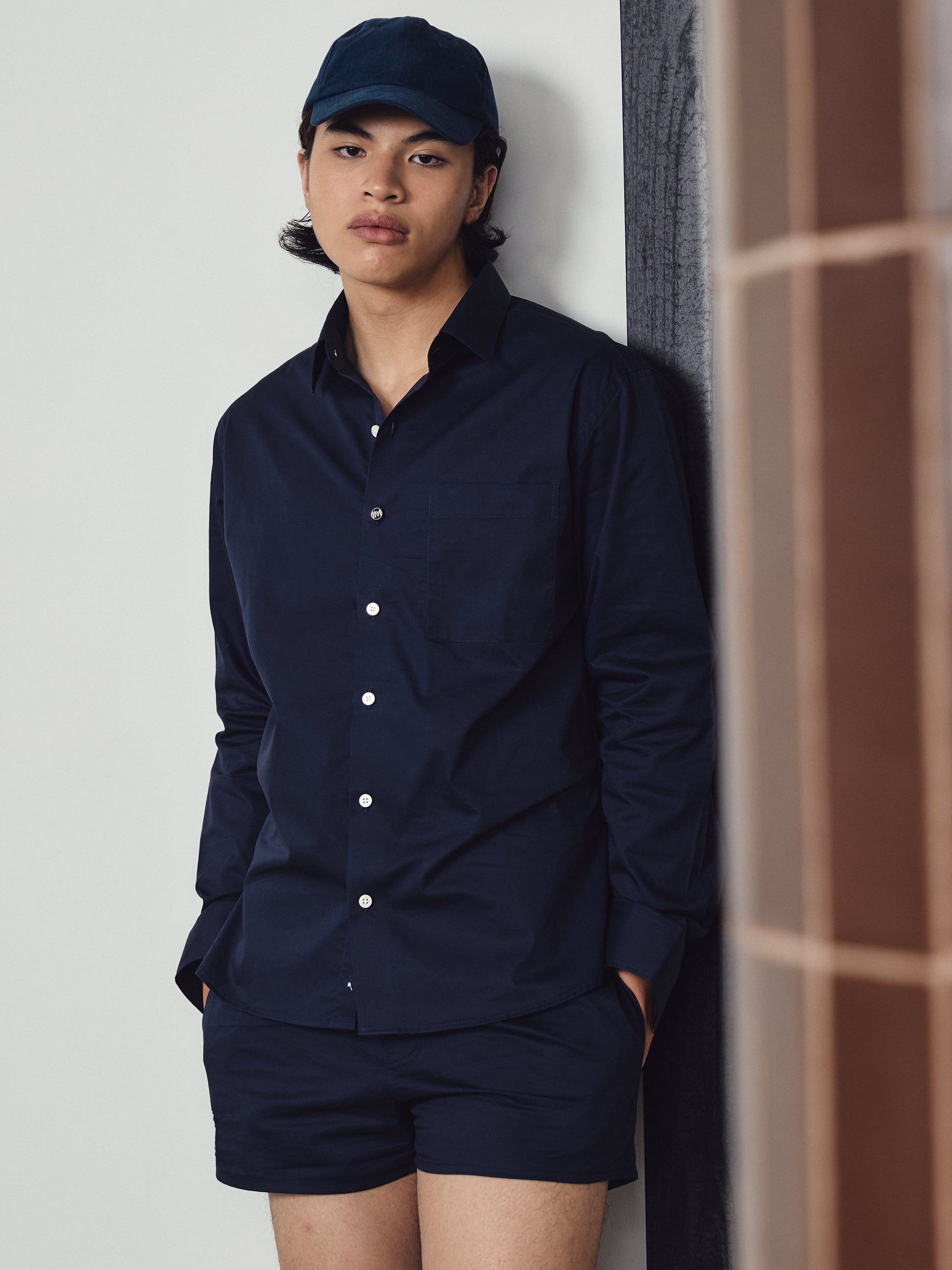 Person wearing a navy shirt and shorts with a cap against a light grey wall.