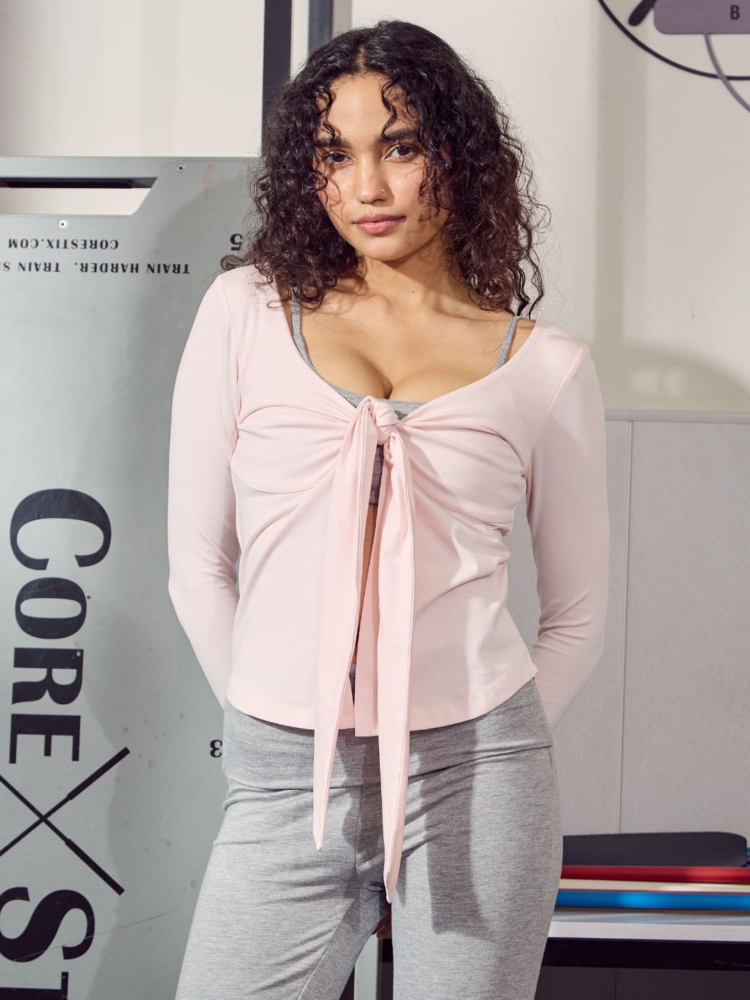 Woman wearing a pink long-sleeve top with a knot detail and grey leggings in front of a Core Strength sign.