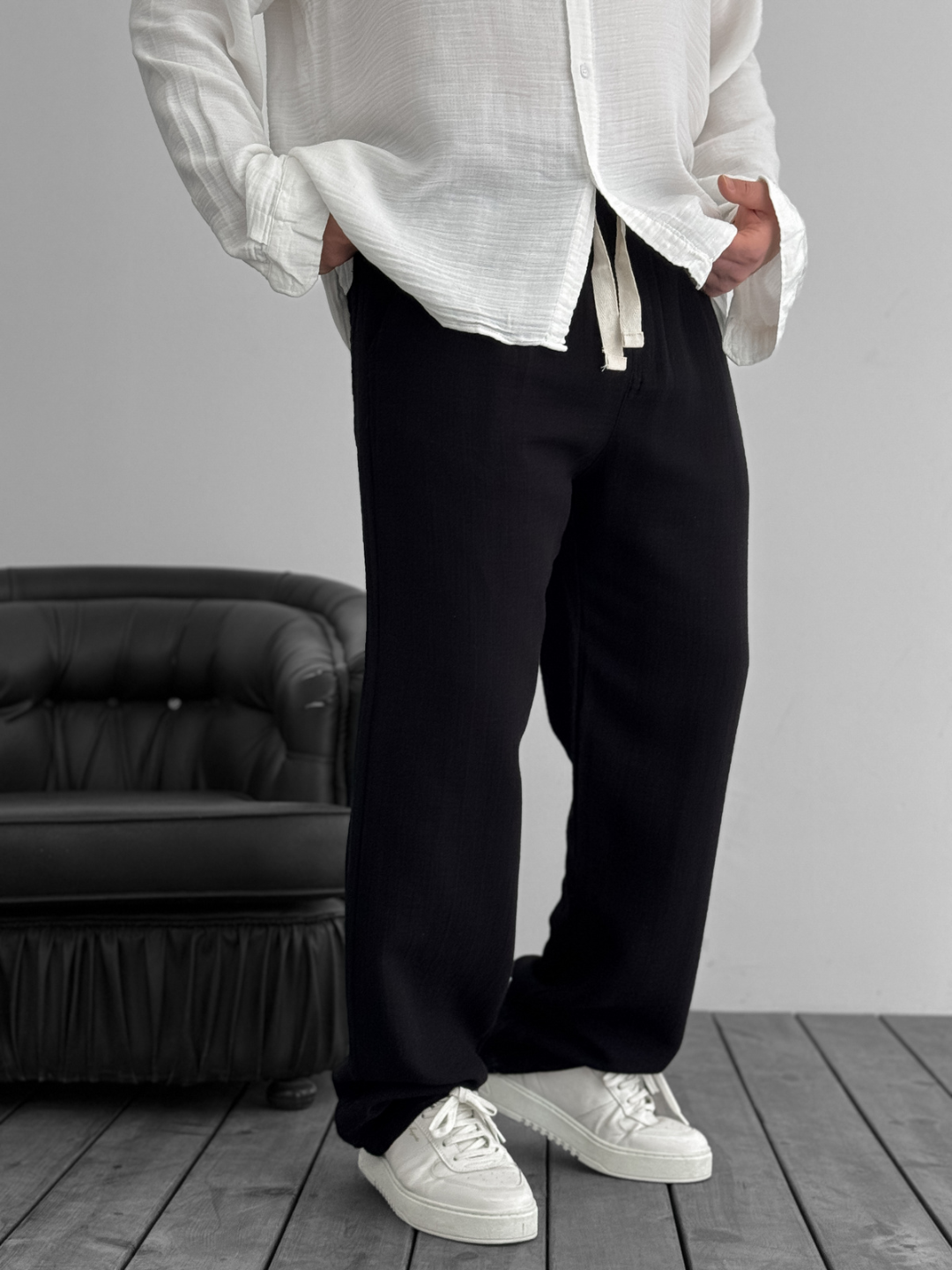 A person standing against a white background, wearing wide-leg black gauze pants and a white shirt with the sleeves rolled up to the elbows.