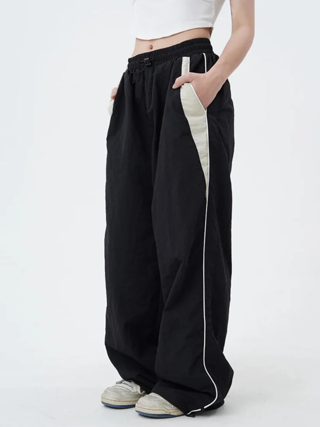 A pair of black track pants with contrasting white piping on the sides, worn by a person standing against a white background.