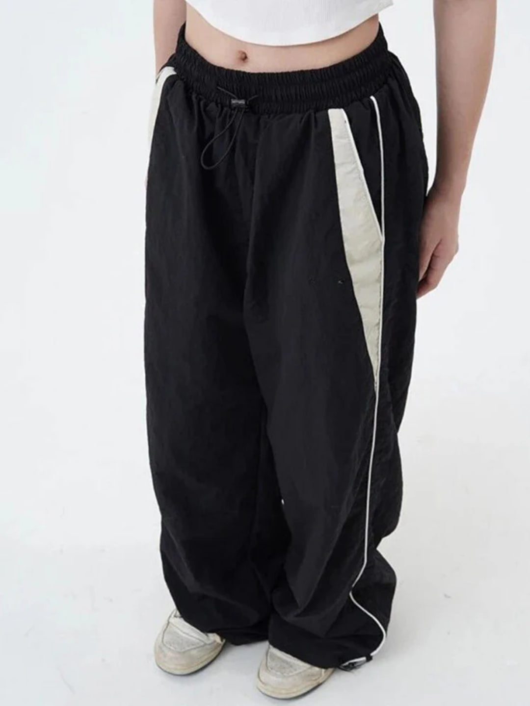 A pair of black track pants with contrasting white piping on the sides, worn by a person standing against a white background.