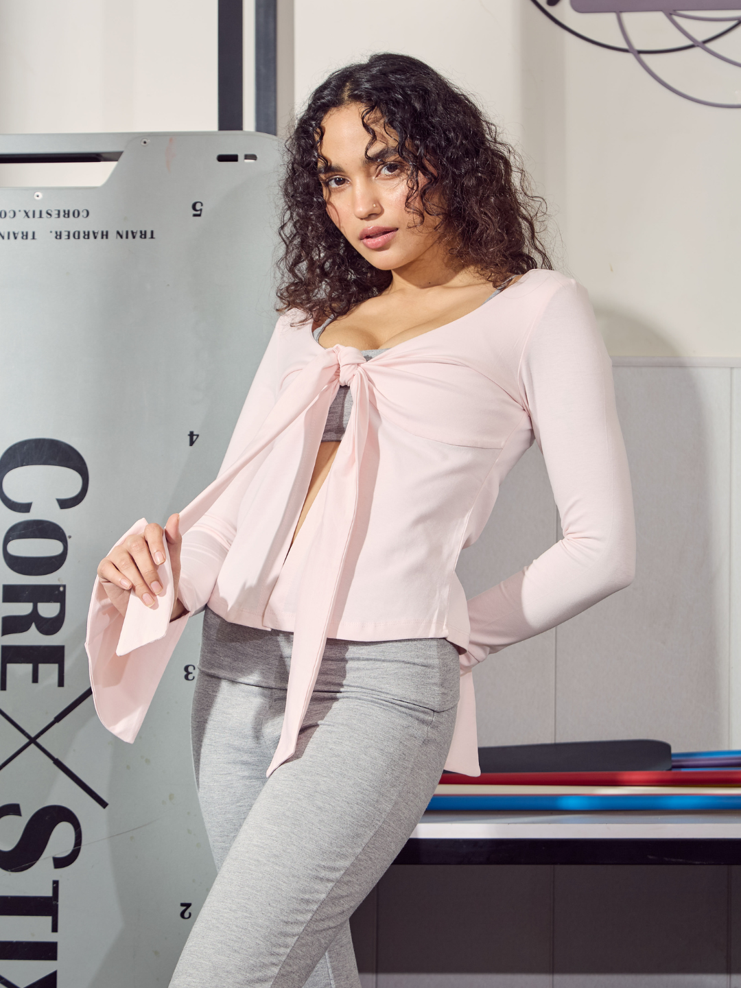 Woman wearing a pink top with a bow and grey pants in front of a white wall with text.