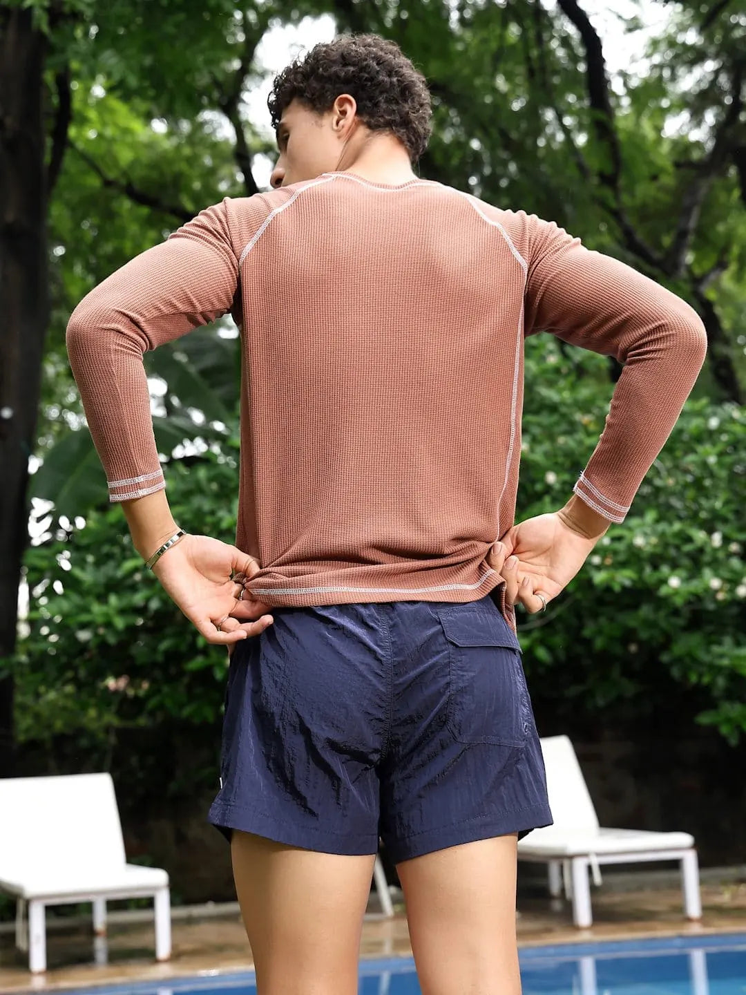 Person wearing a long-sleeve brown shirt and navy shorts standing by a pool with greenery in the background.