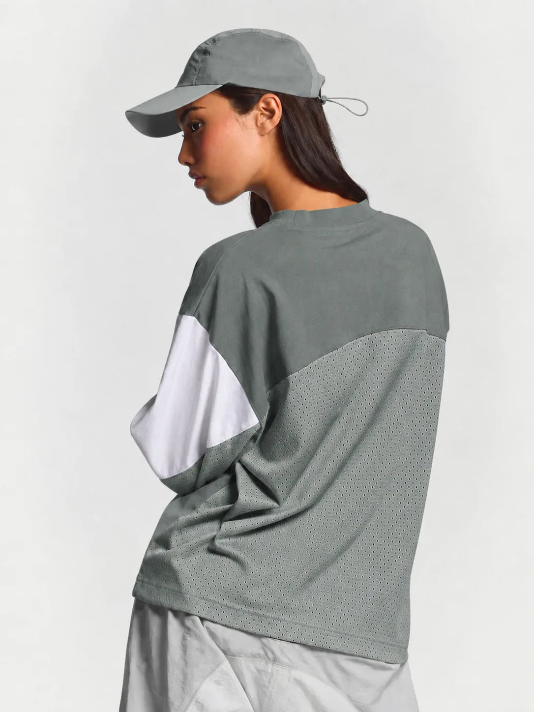 A woman wearing a grey and white color-blocked long-sleeve athletic top, a grey baseball cap, and light grey shorts