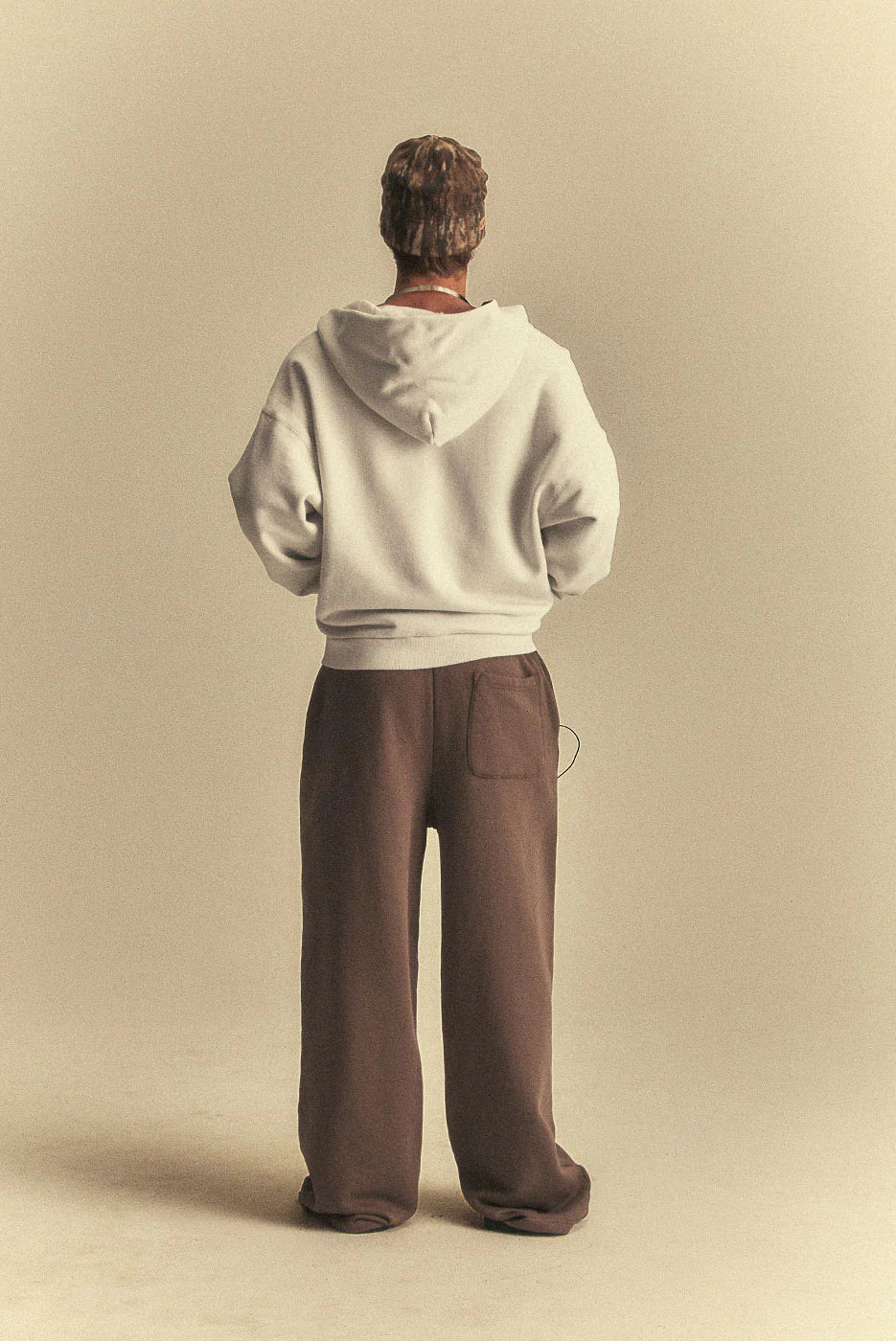 Person wearing a light grey hoodie and brown pants standing against a beige background