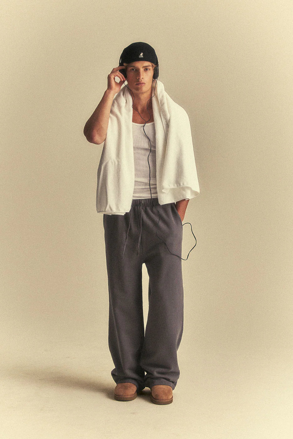 Man wearing a cap, white shirt, grey pants, and brown shoes on a beige background