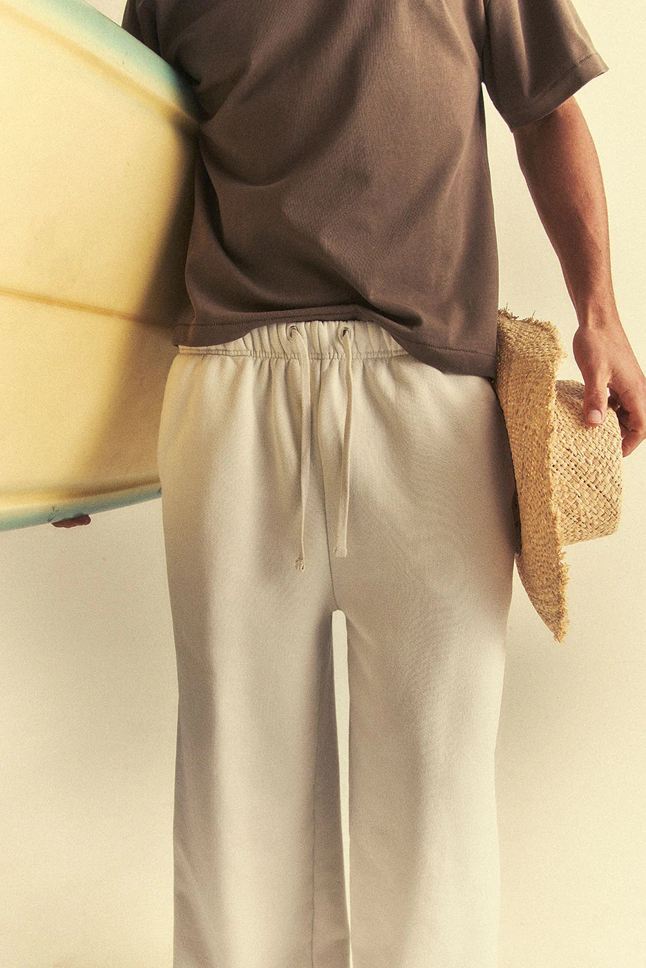 Person holding a surfboard and a hat, wearing a brown shirt and beige pants, against a neutral background.
