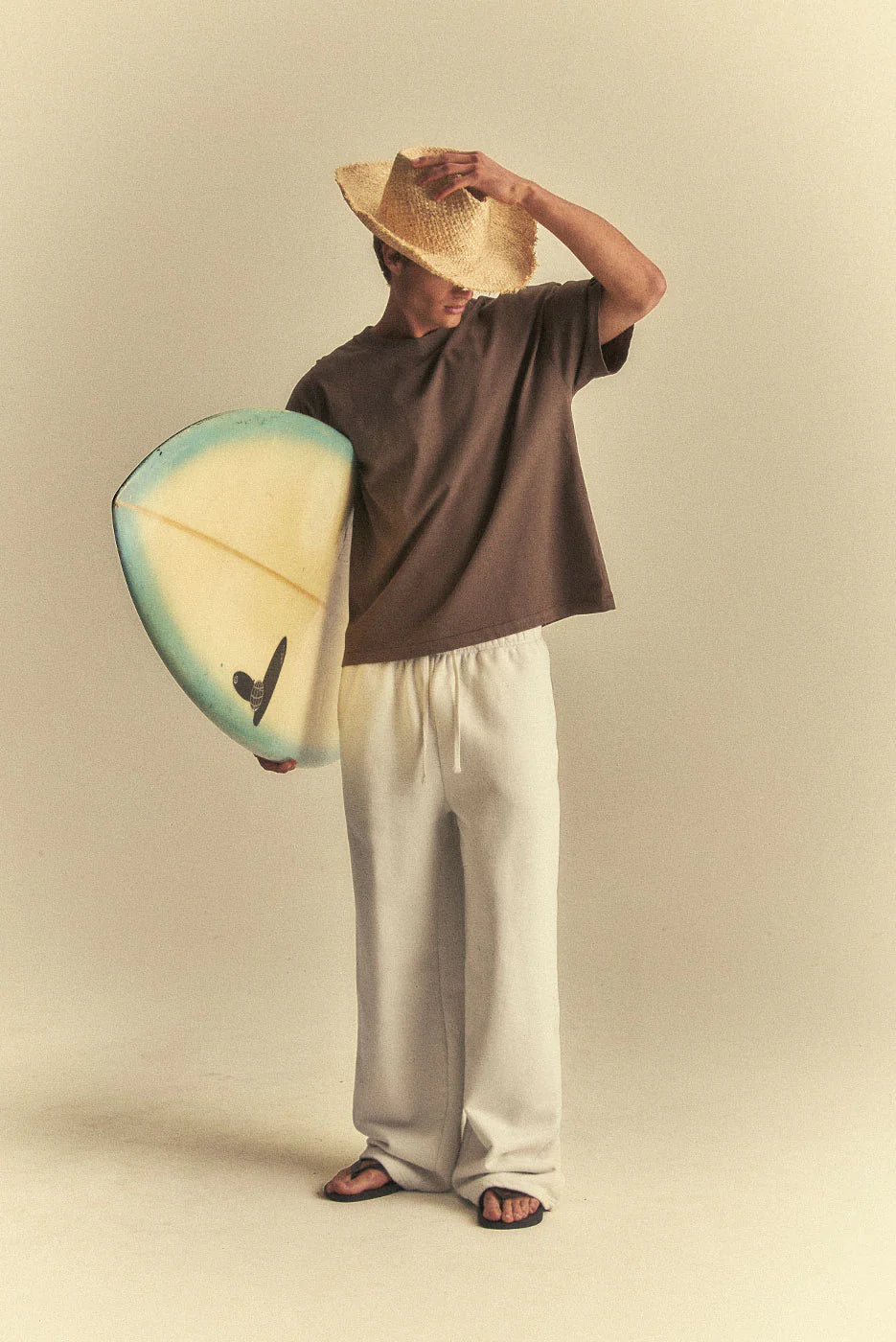 Person holding a surfboard and wearing a hat against a beige background
