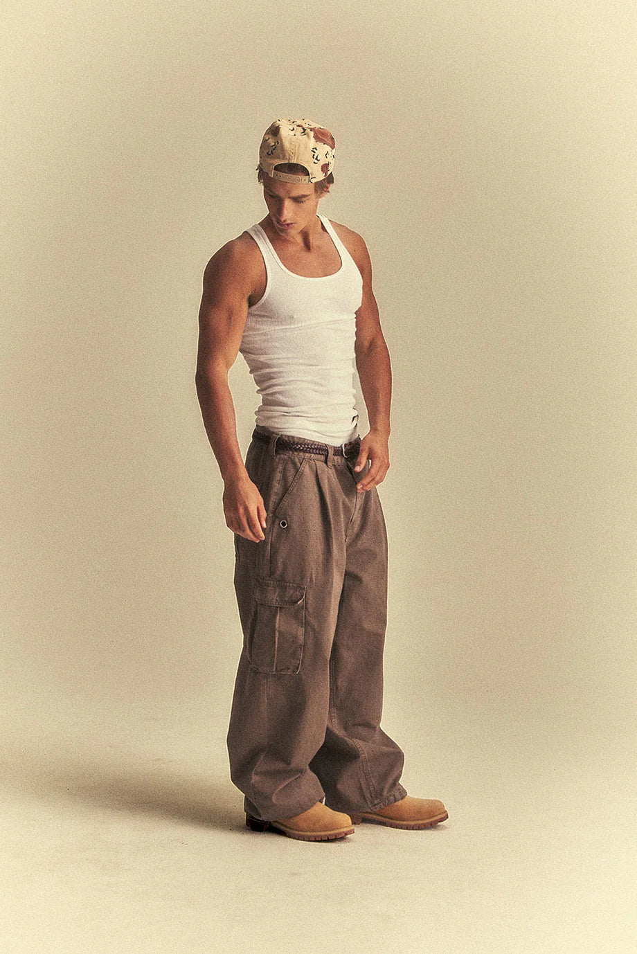 Man wearing a white tank top, brown pants, and a cap on a beige background