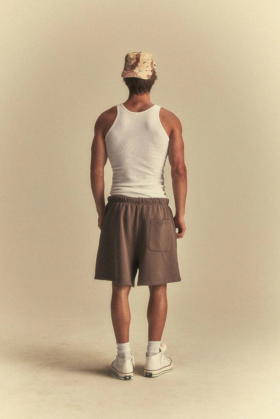 Person wearing a white tank top, brown shorts, and a beige bucket hat on a beige background