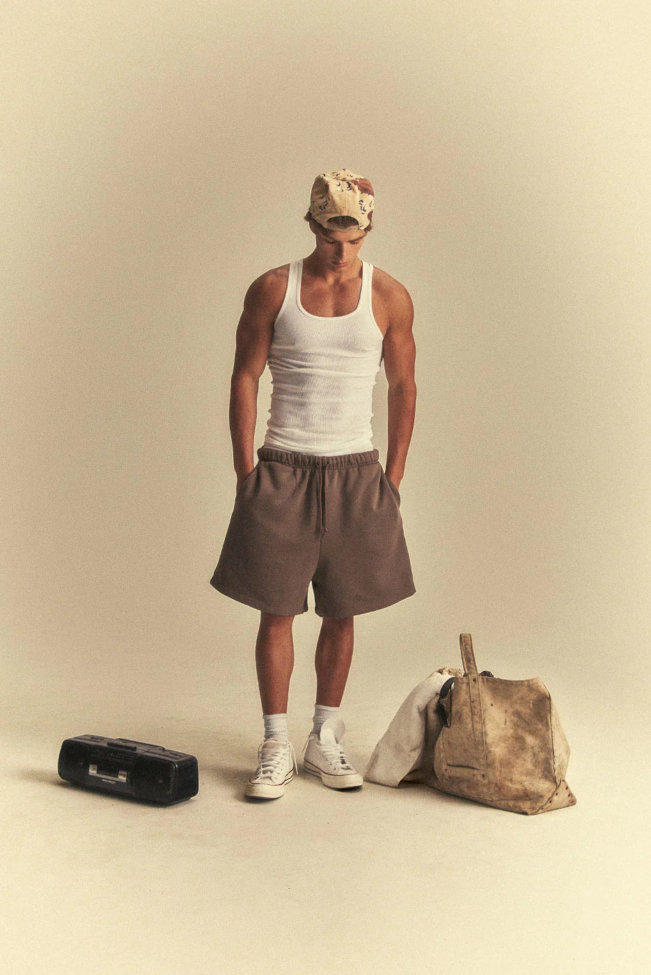 Person wearing a white tank top and brown shorts standing next to a bag and radio on a beige background