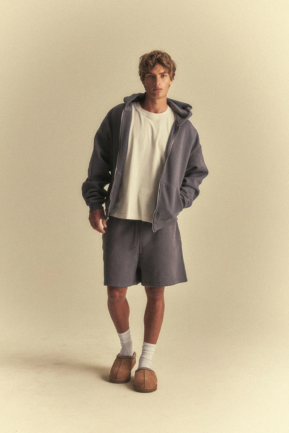 Person wearing a grey hoodie, white t-shirt, grey shorts, and brown shoes on a beige background