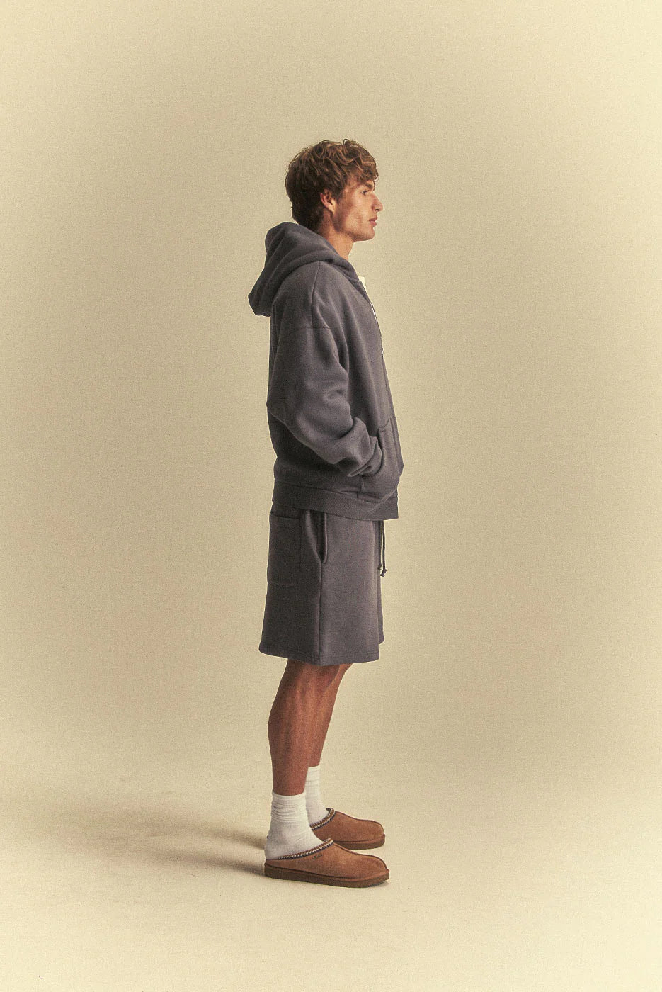 Man wearing a grey hoodie and shorts standing against a beige background