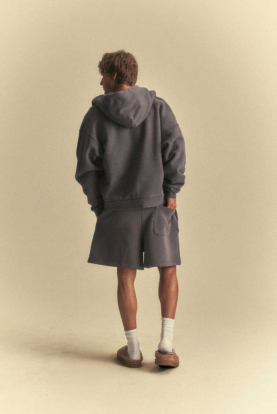 Person wearing a grey hoodie and shorts standing against a beige background
