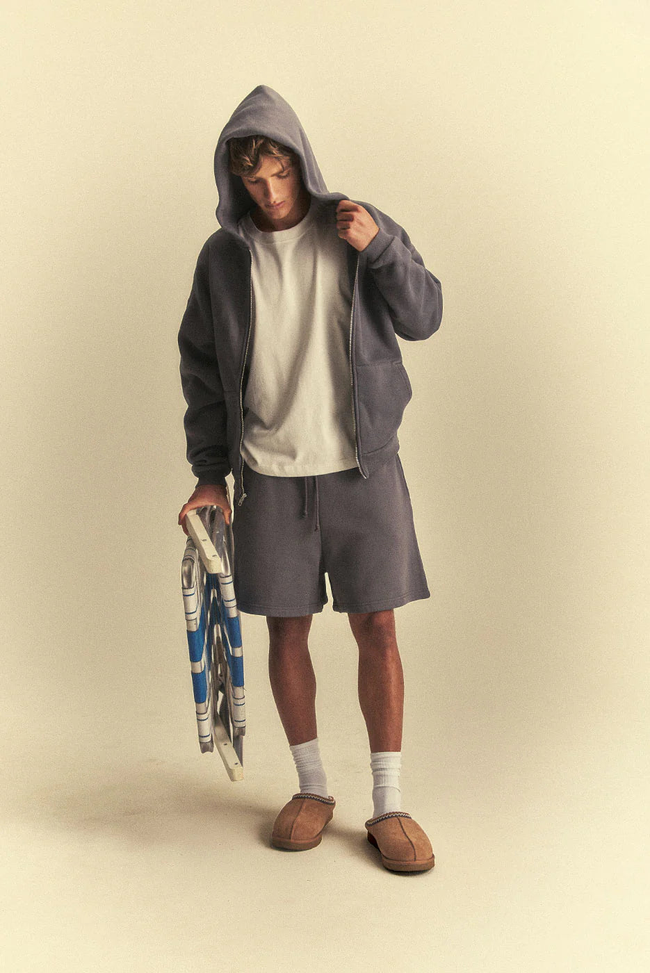 Person wearing a grey hoodie and shorts holding a tennis racket on a beige background