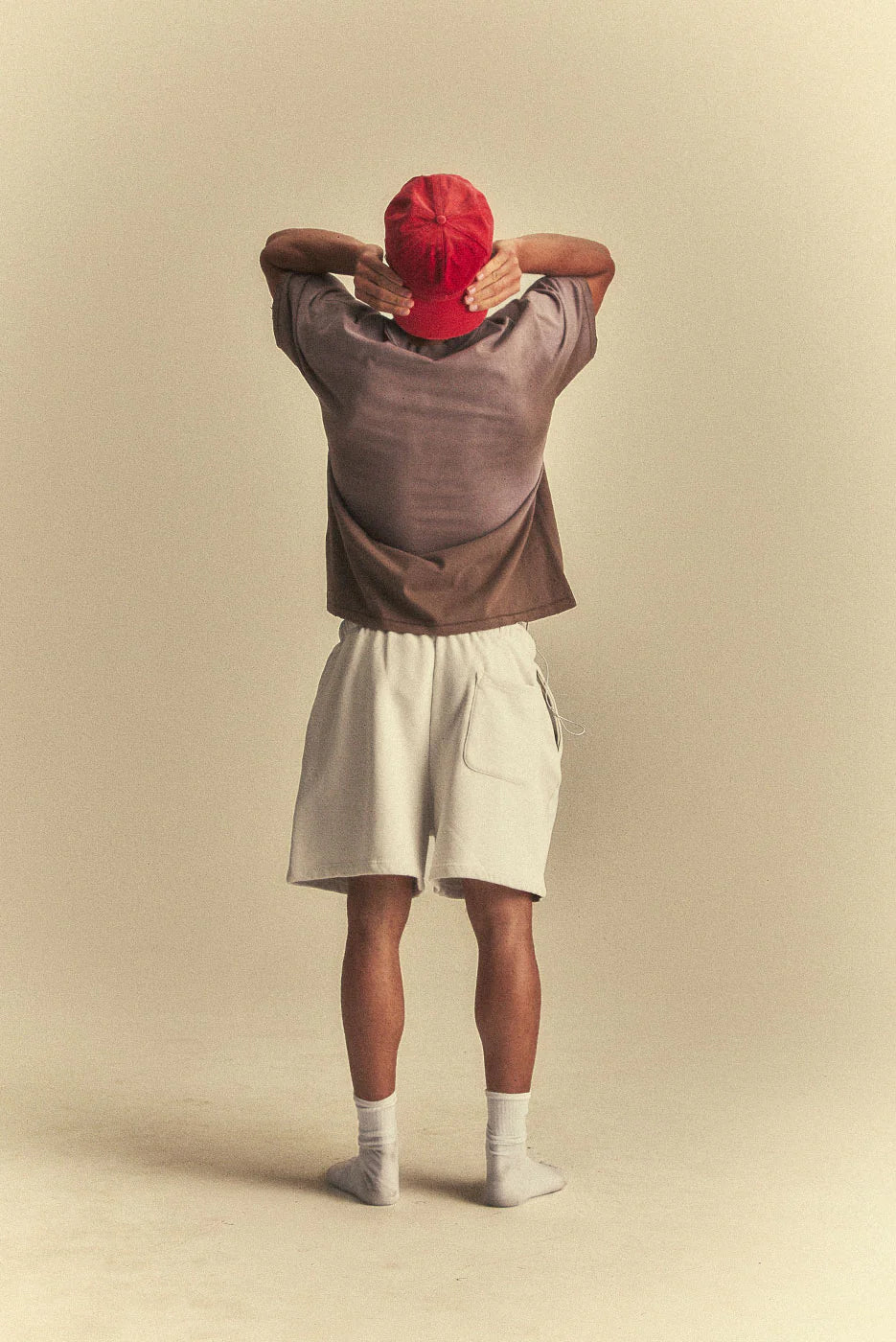 Person wearing a red cap, brown sweater, and white shorts against a beige background