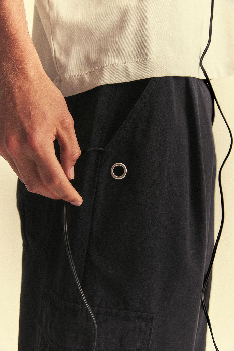 Person wearing black pants with a button, holding a black cord against a beige background