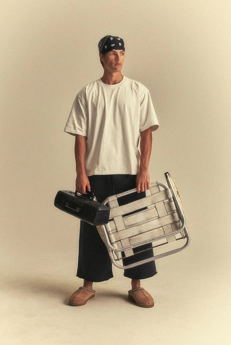 Person holding a black suitcase and a metal folding chair against a beige background
