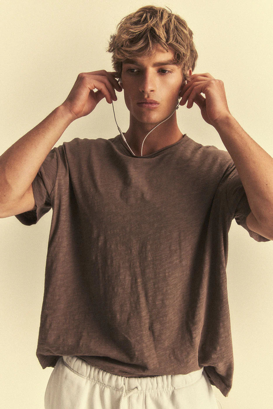 Man wearing a brown t-shirt and white pants, adjusting earphones against a beige background