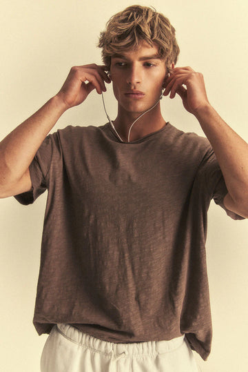 Man wearing a brown t-shirt and white pants, adjusting earphones against a beige background