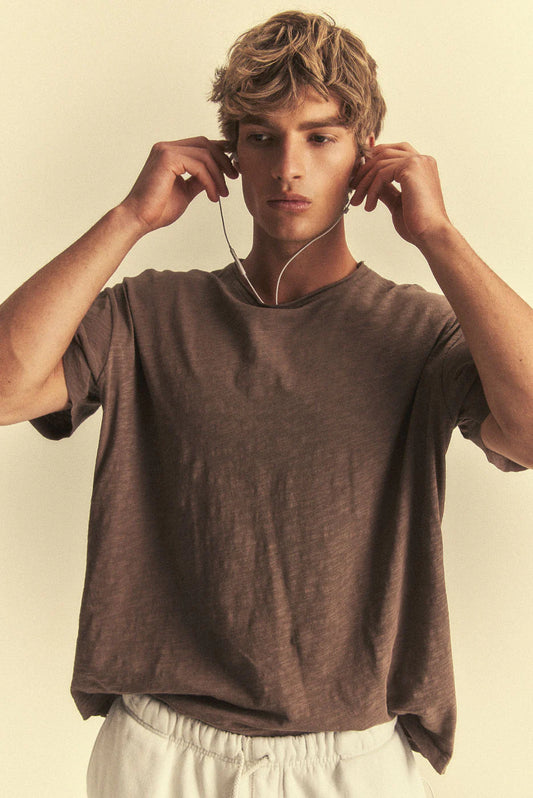 Man wearing a brown t-shirt and white pants, adjusting earphones against a beige background
