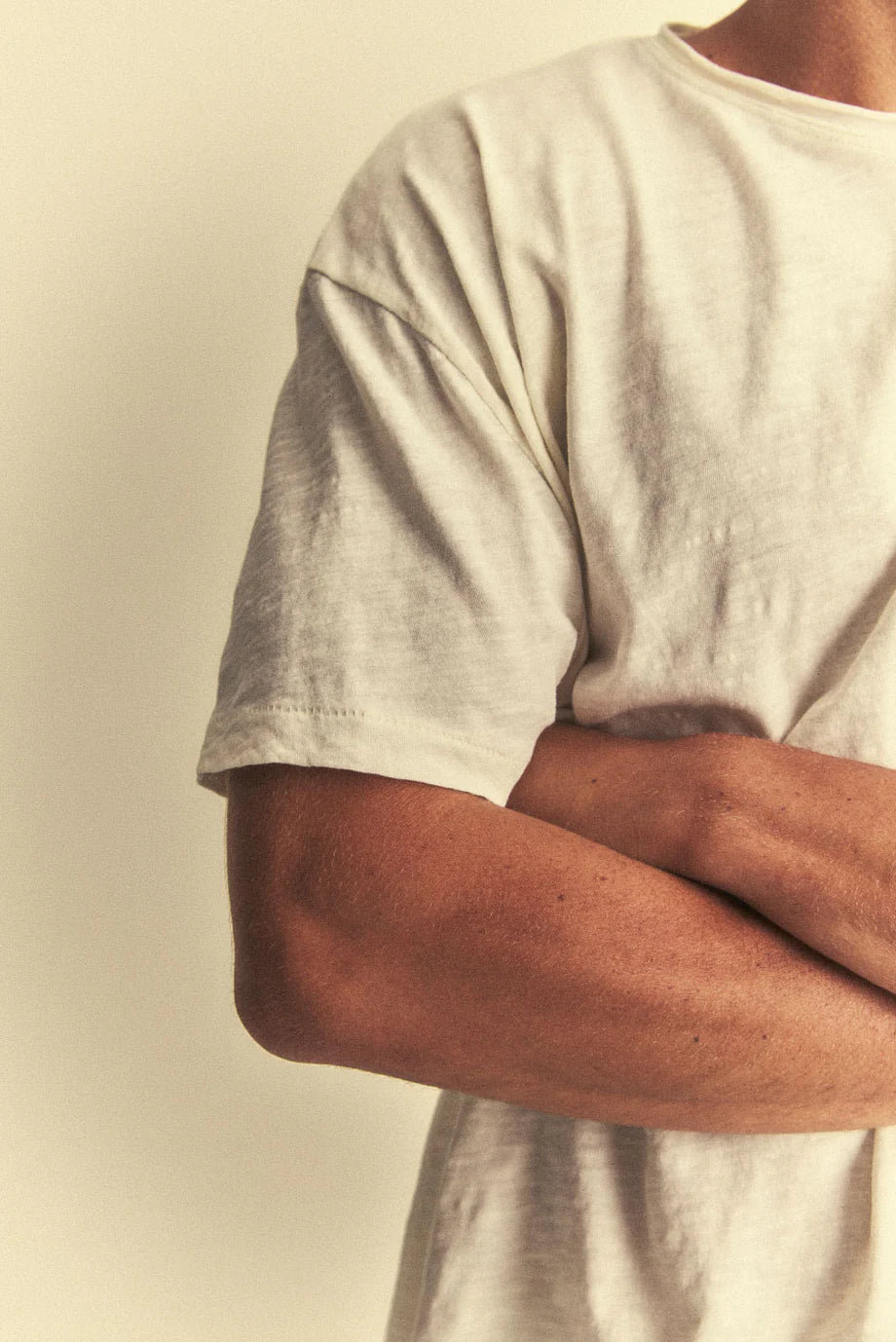 Person wearing a plain white t-shirt with arms crossed on a beige background