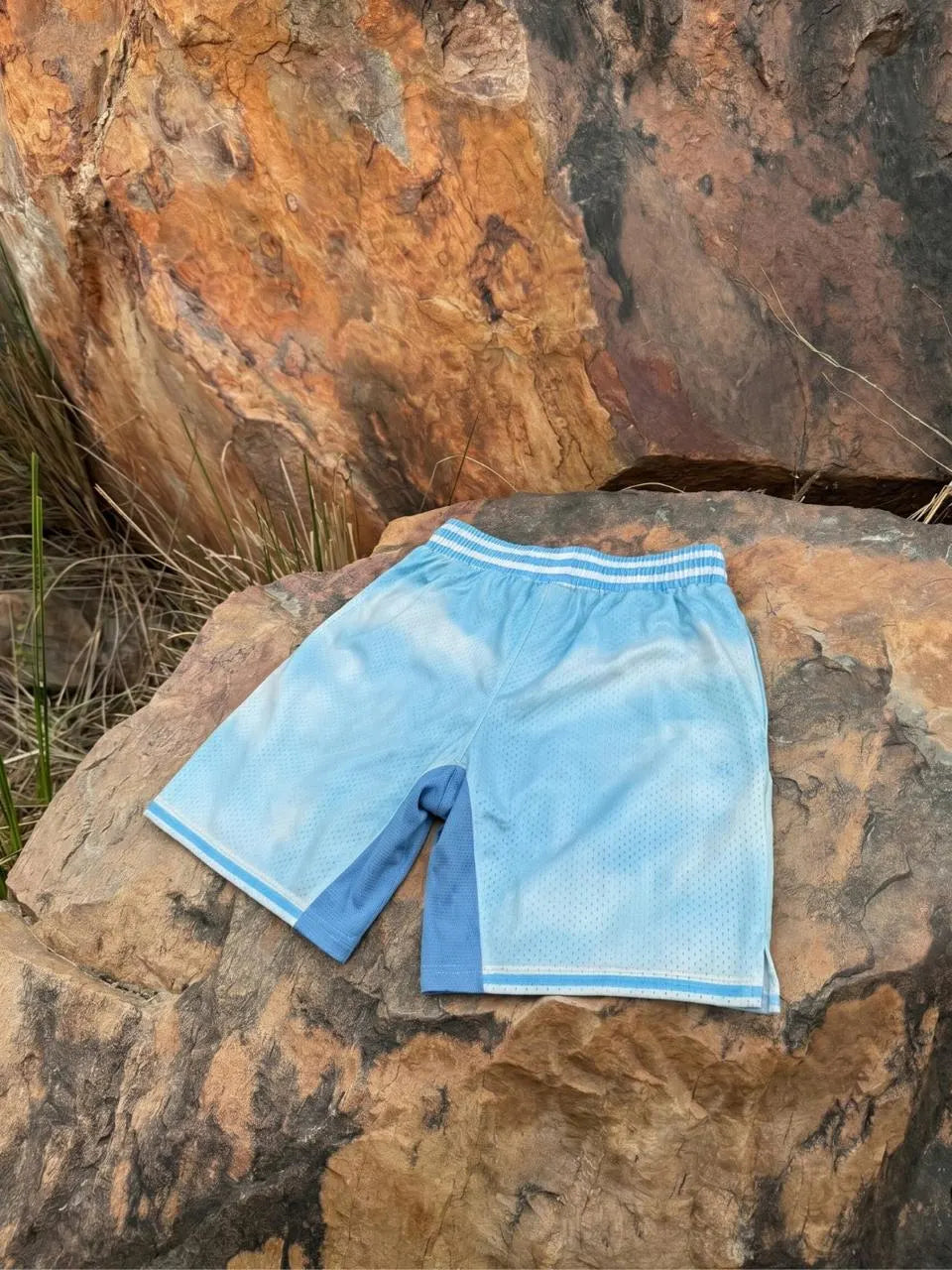 Light blue athletic shorts with a white striped waistband and white trim, laid flat on a large brown rock outdoors.
