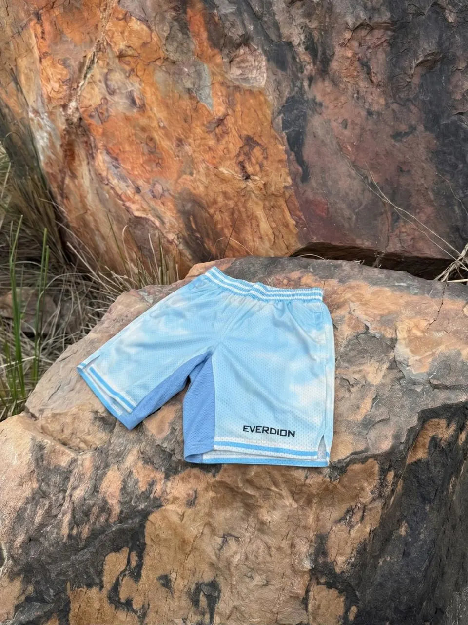 Light blue athletic shorts with white trim and the brand name 'EVERDION' printed in black near the hem, displayed on a large brown rock outdoors.