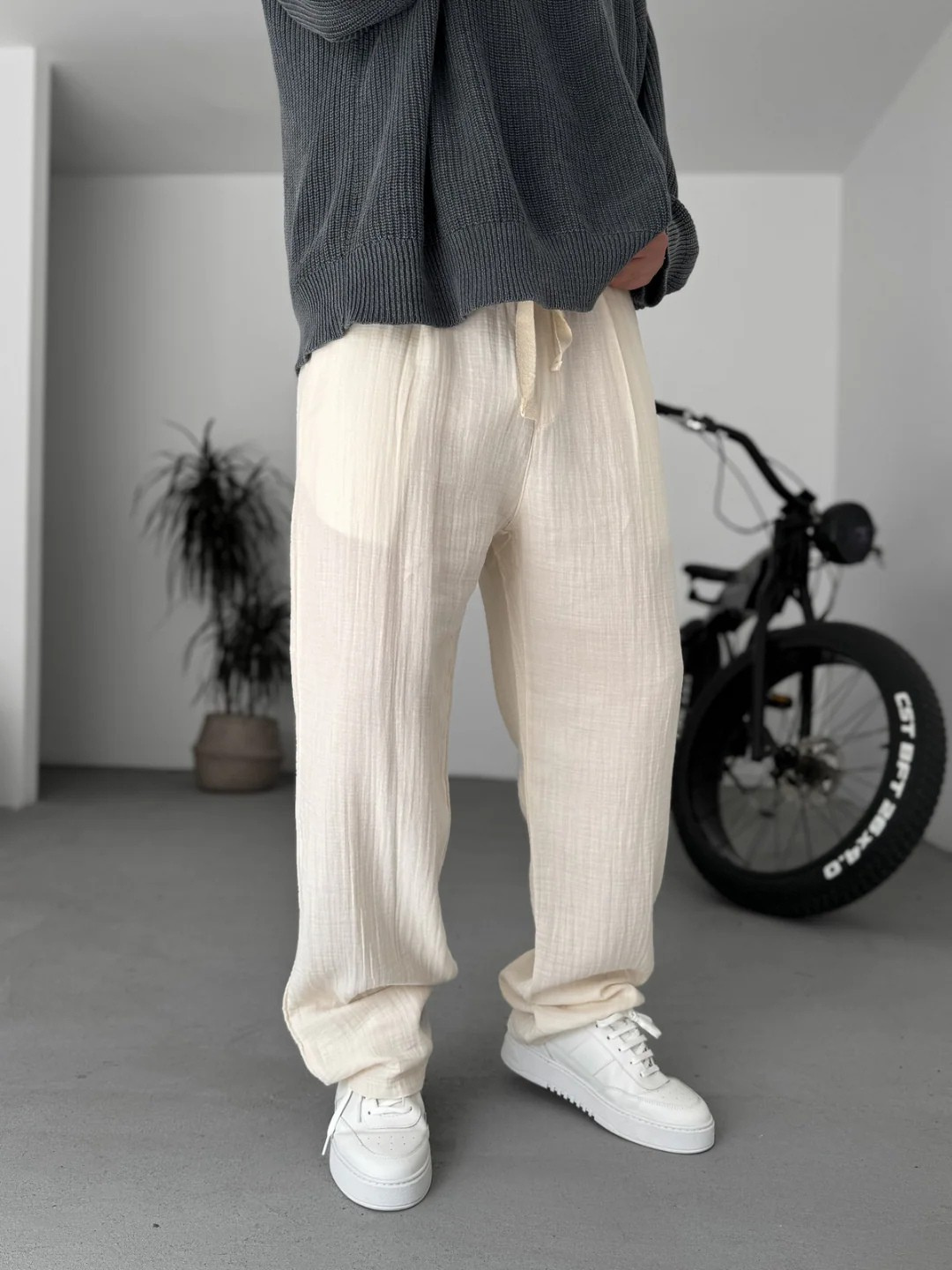 A person standing against a white background, wearing wide-leg gray gauze pants and a white shirt with the sleeves rolled up to the elbows.