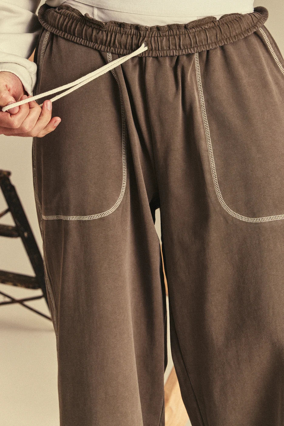 Brown sweatpants with white stitching detail worn by a person, held up to show fit.