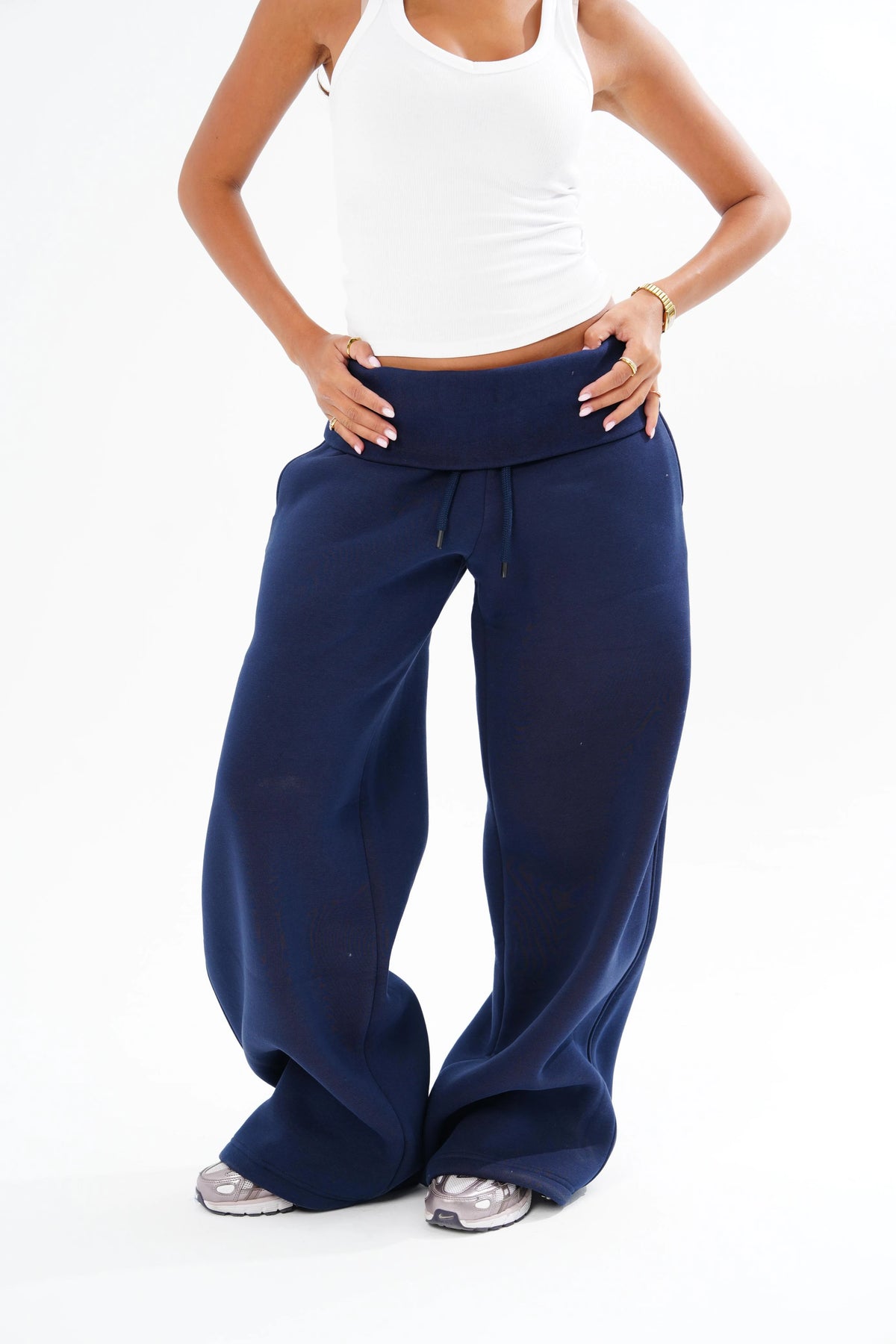 A model wearing navy blue wide-leg sweatpants with a drawstring waistband, paired with a white tank top and grey sneakers, shown from the chest down on a white studio background.