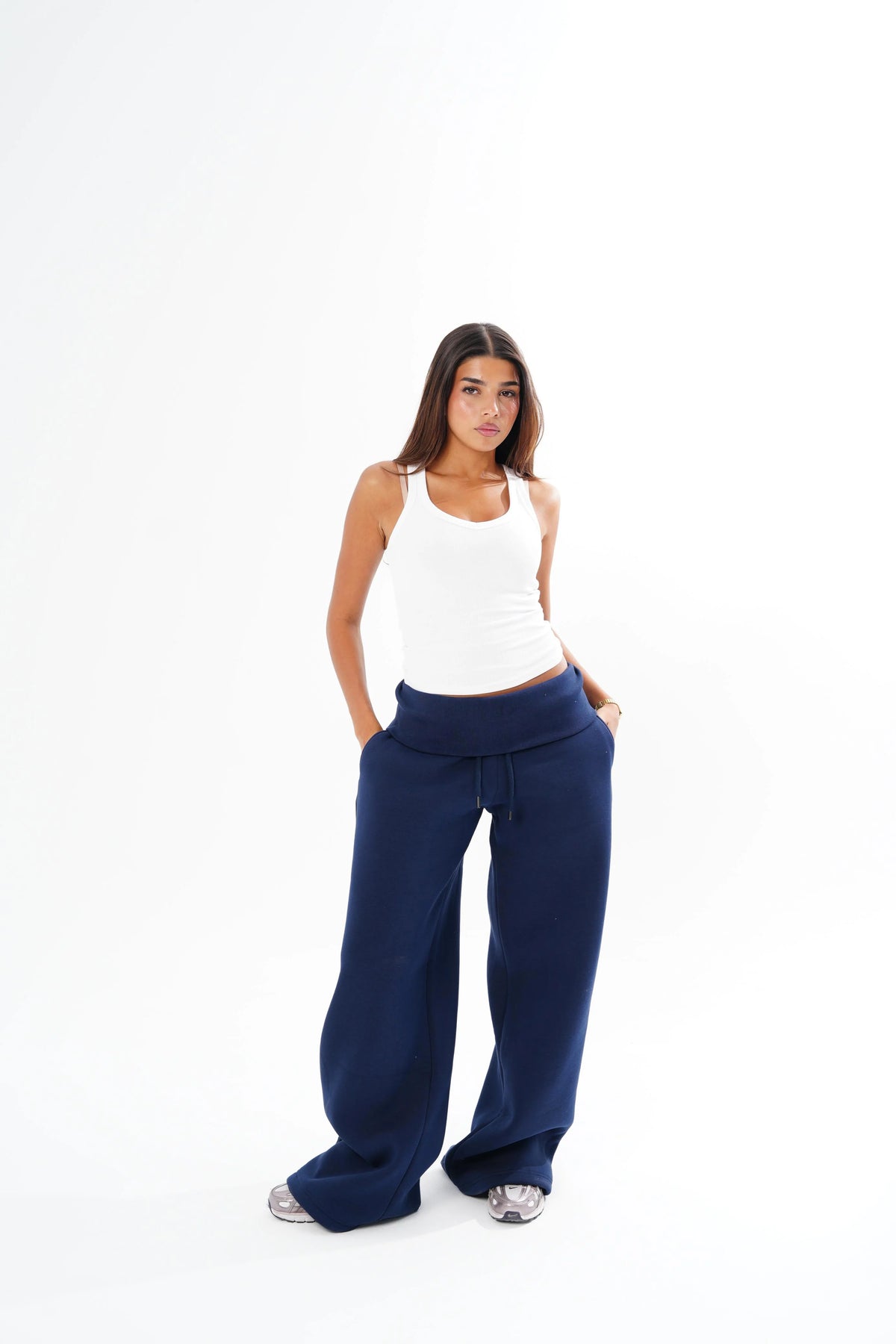A woman modeling a pair of wide-leg navy blue sweatpants with a white ribbed tank top, standing against a solid white studio background.