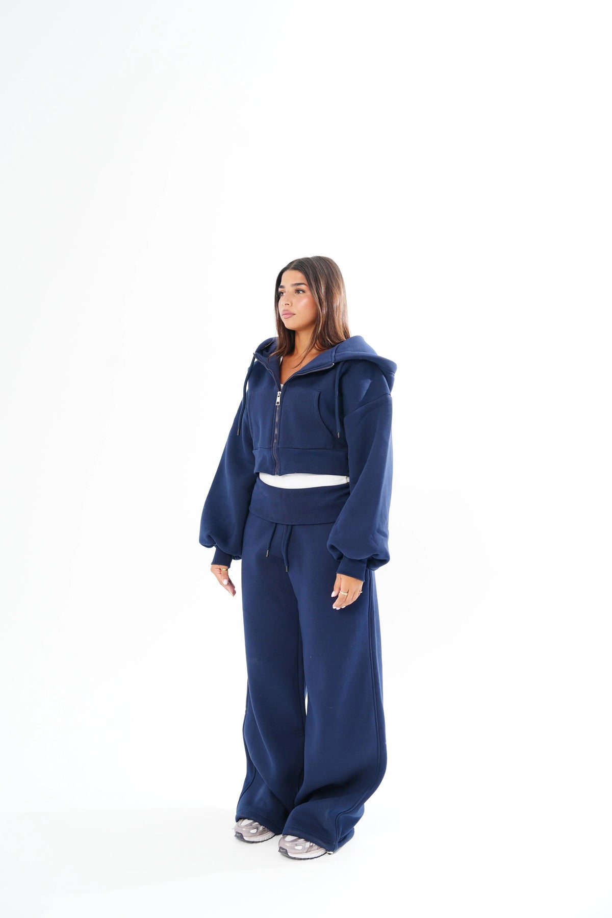 A woman modeling a matching navy blue lounge set consisting of a cropped zip-up hoodie with long sleeves and wide-leg sweatpants, standing against a solid white background.
