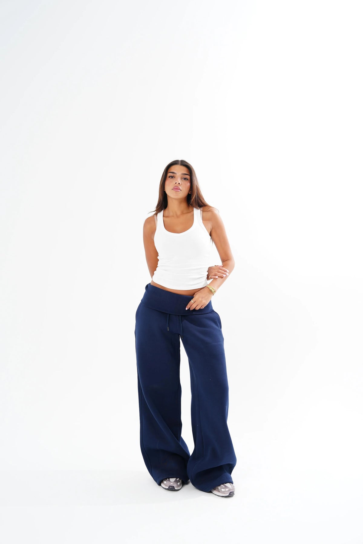 A woman modeling a pair of wide-leg navy blue sweatpants with a white ribbed tank top, standing against a solid white studio background.