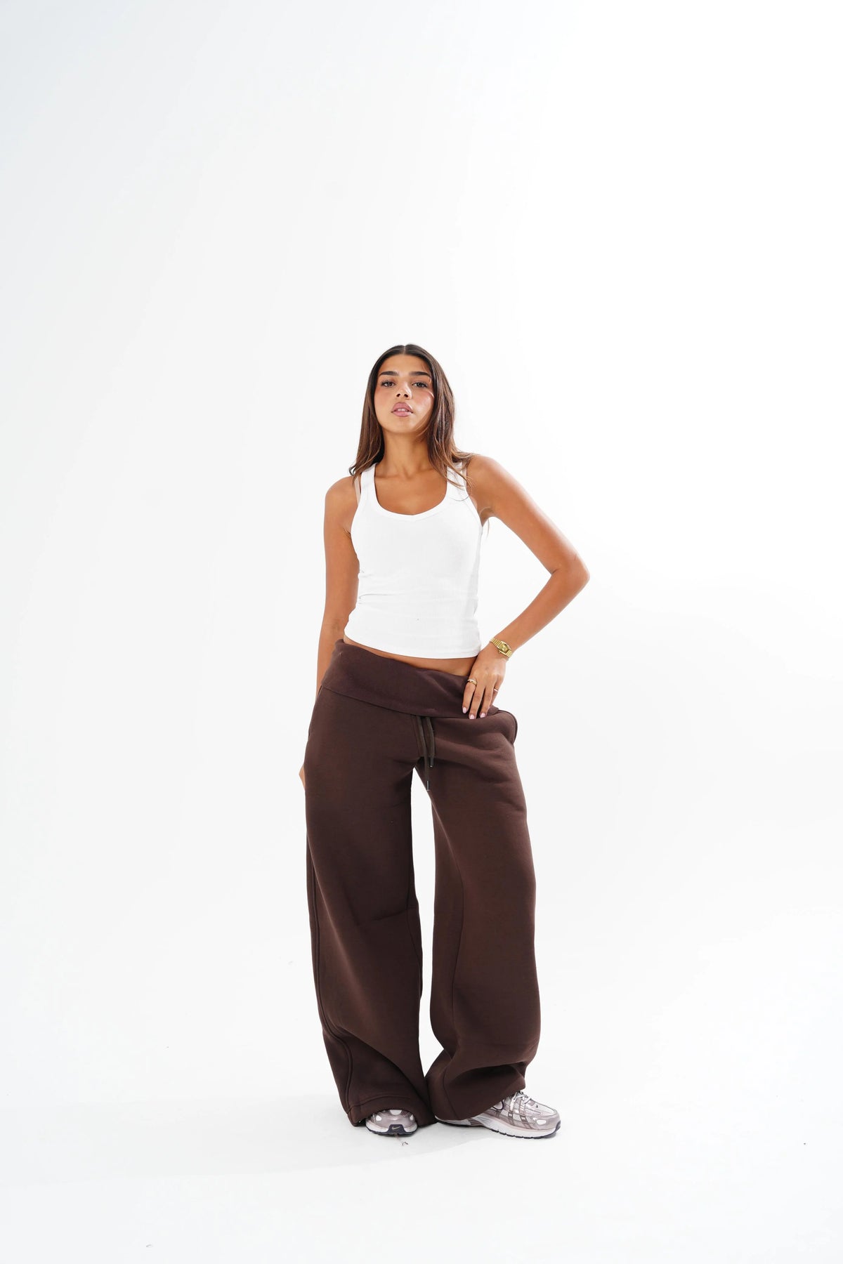 A woman modeling a pair of wide-leg brown sweatpants with a white ribbed tank top, standing against a solid white studio background.