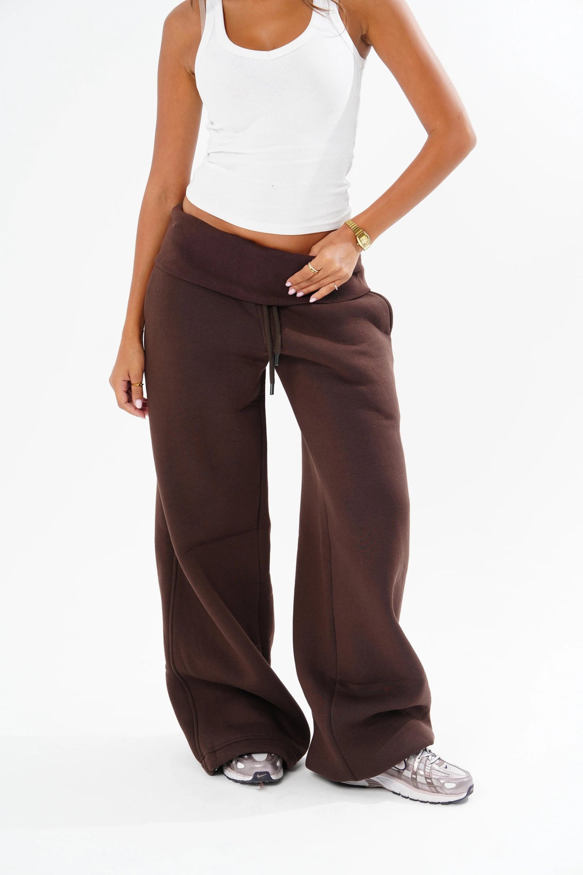 A model wearing a pair of wide-leg brown sweatpants with a drawstring waist, styled with a white tank top and grey sneakers against a plain white background.