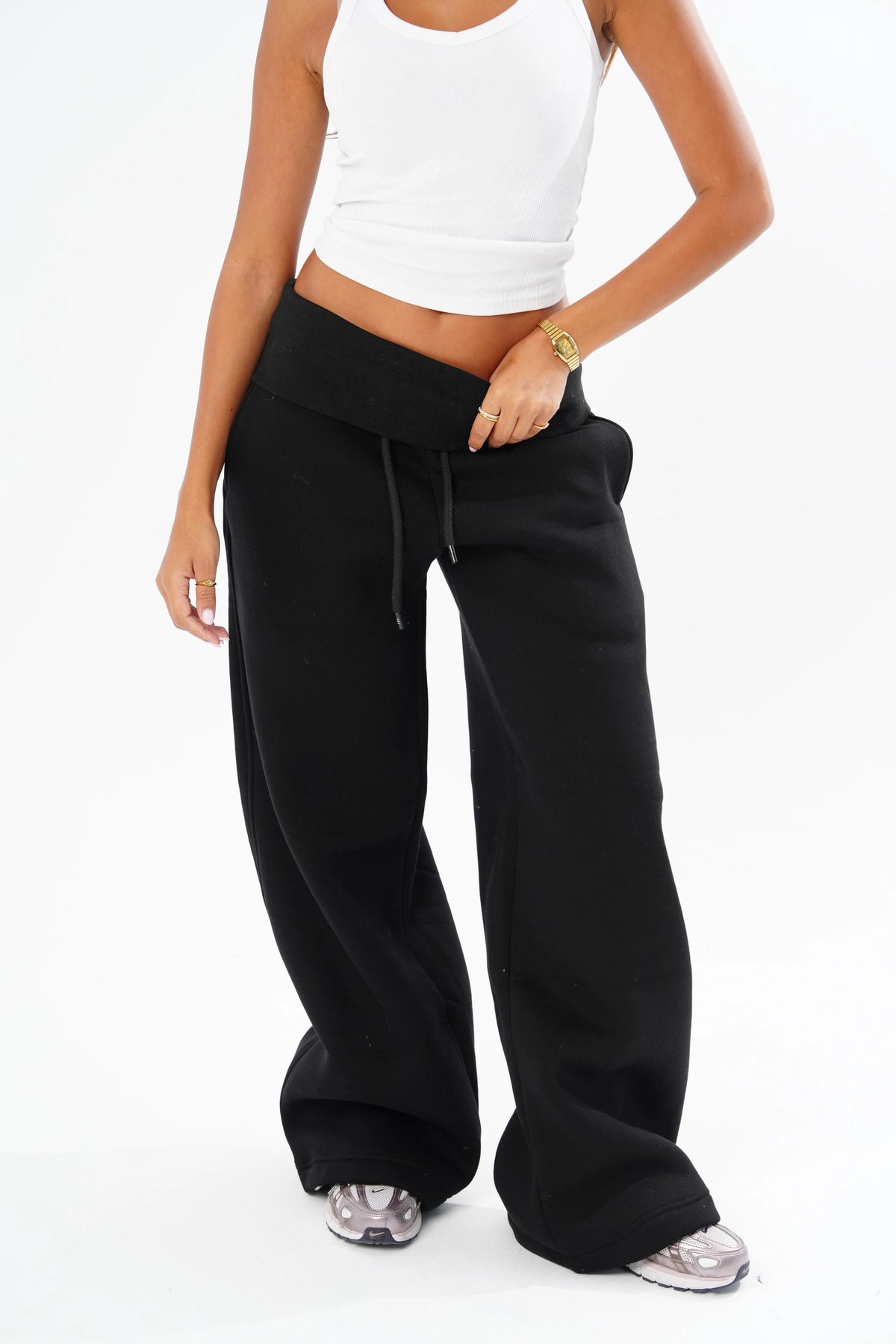 A model wearing black wide-leg sweatpants with a drawstring waistband, styled with a white crop top and silver sneakers against a white studio background.