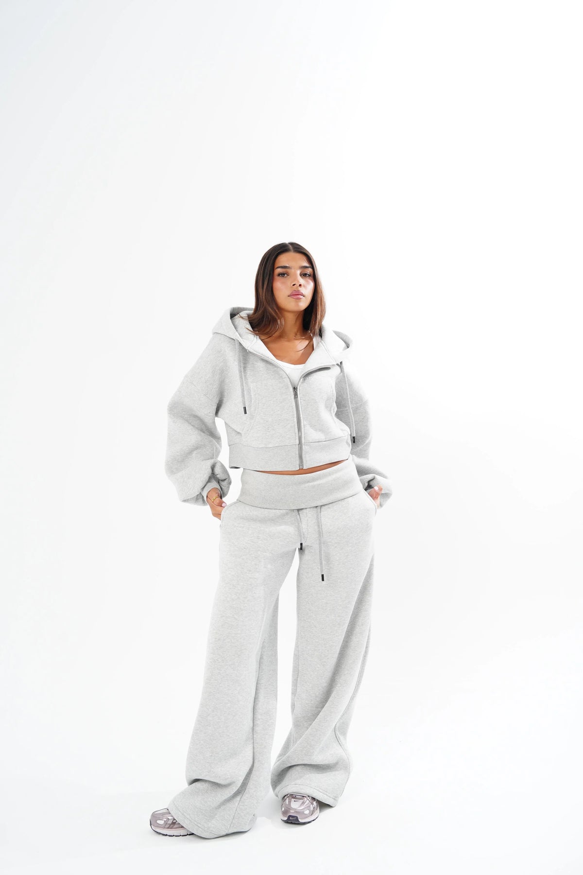 A woman modeling a light grey two-piece lounge set consisting of a cropped zip-up hoodie and matching wide-leg sweatpants, photographed against a seamless white studio background.