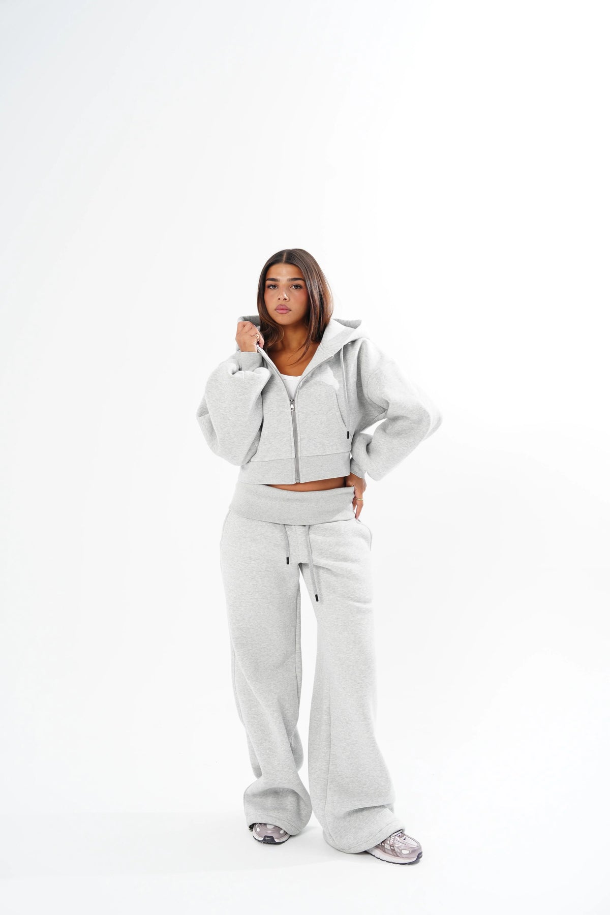 A woman modeling a light grey matching lounge set consisting of a cropped zip-up hoodie and wide-leg sweatpants, standing against a solid white studio background.