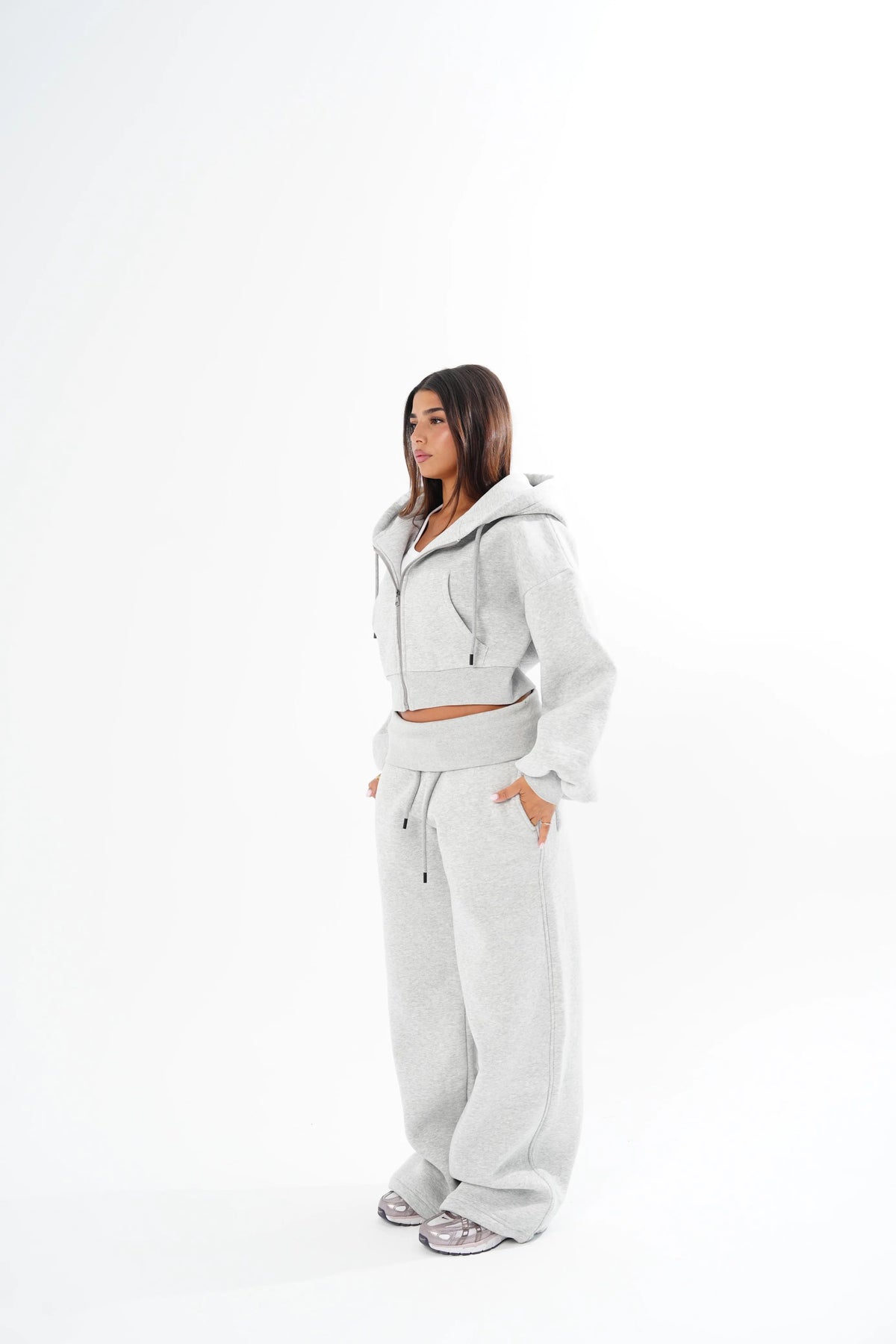A woman modeling a light grey two-piece lounge set consisting of a cropped zip-up hoodie and matching wide-leg sweatpants, standing against a solid white studio background.