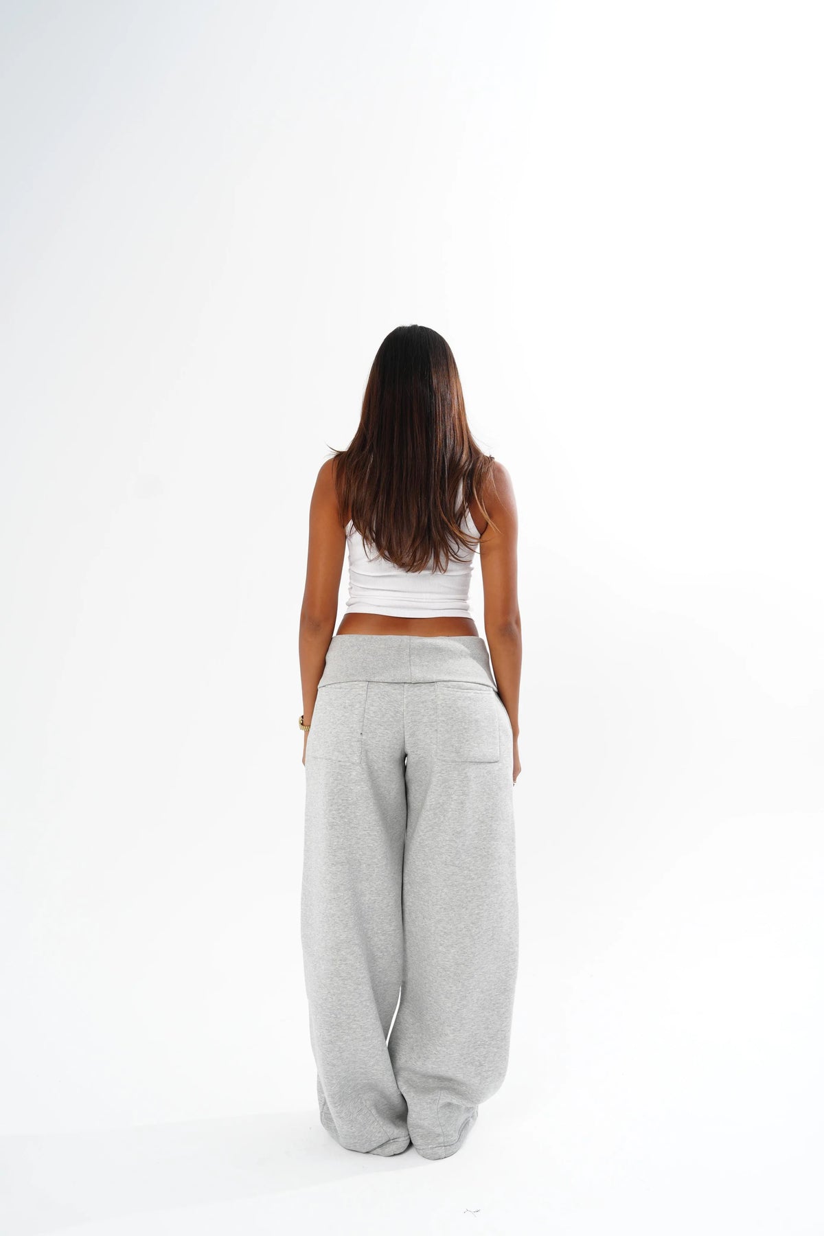A woman viewed from behind wearing light grey wide-leg sweatpants and a white cropped tank top, standing against a plain white studio background.