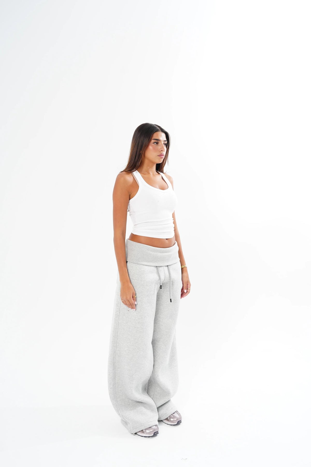 A woman stands in profile wearing light grey wide-leg sweatpants, a white ribbed tank top, and gray sneakers against a plain white studio background.
